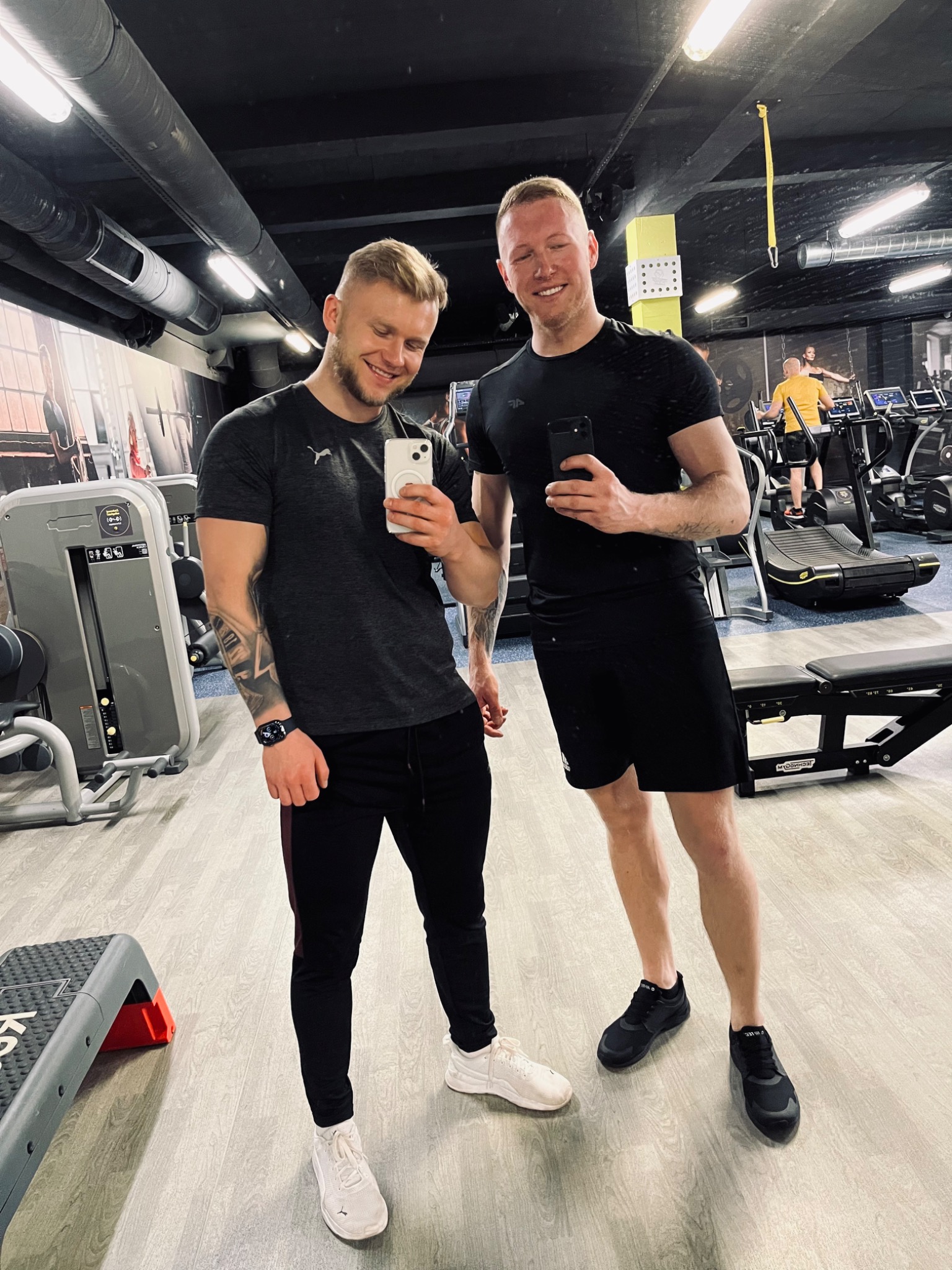 Dwaj uśmiechnięci mężczyźni robią selfie w siłowni, w tle widoczne maszyny do ćwiczeń i osoby trenujące.