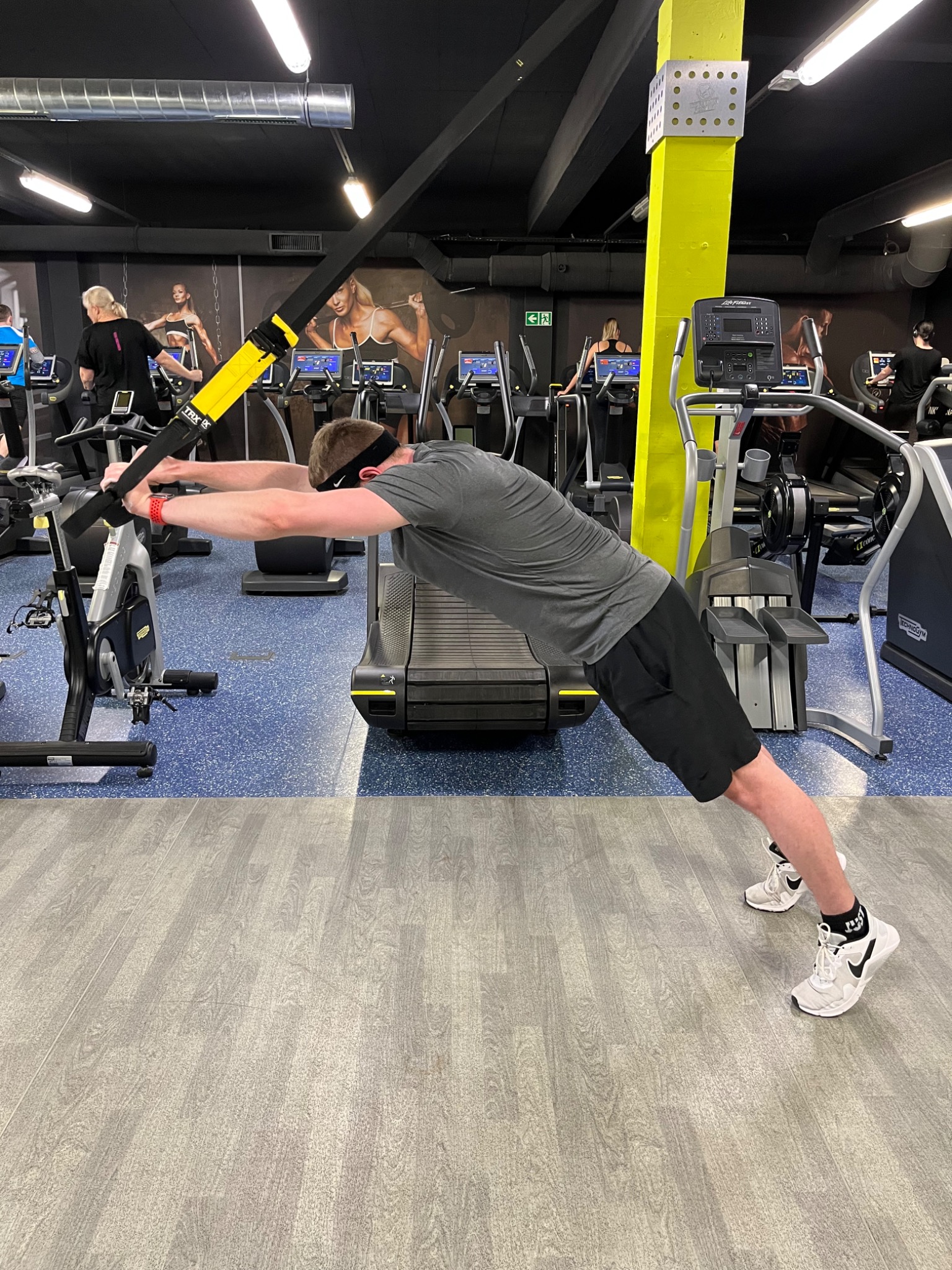 Mężczyzna w szarej koszulce i czarnych spodenkach wykonuje ćwiczenie rozciągające na TRX w siłowni, z widocznym bieżnią, rowerem stacjonarnym i innymi osobami ćwiczącymi w tle.