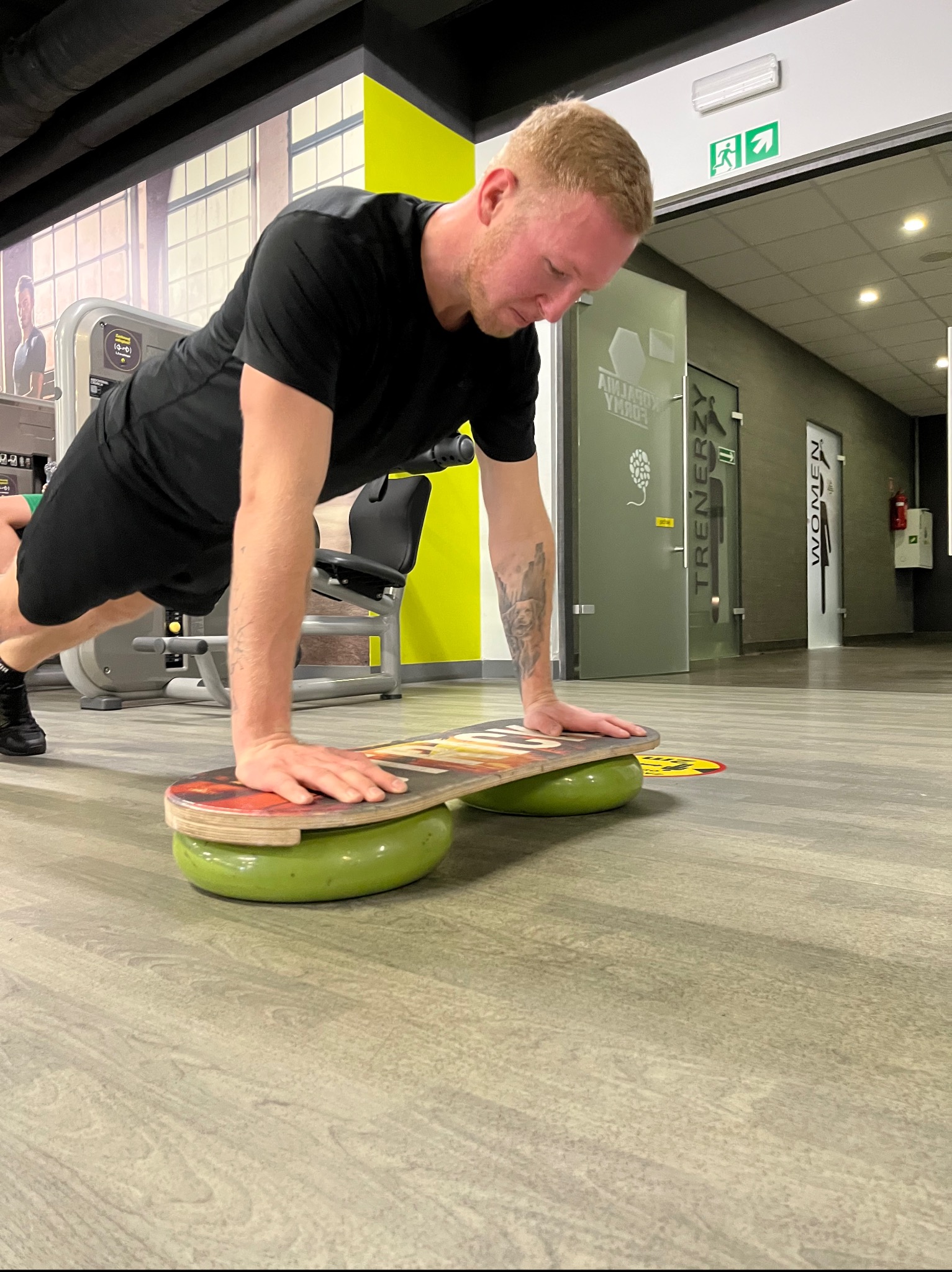 Mężczyzna w Jastrzębiu-Zdroju wykonuje pompkę na desce balansującej w siłowni, z widocznym tatuażem na ramieniu i sprzętem do ćwiczeń w tle.