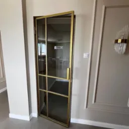 Otwarta, szklana ścianka działowa z złotymi profilami, oddzielająca pomieszczenia w nowoczesnym wnętrzu.