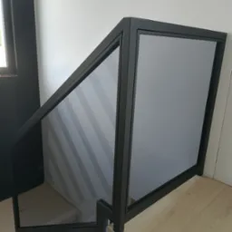 Nowoczesna balustrada szklana z czarną ramą, zamontowana na schodach wewnętrznych. Minimalistyczny design, jasne tło, widoczne schody i fragment podłogi.