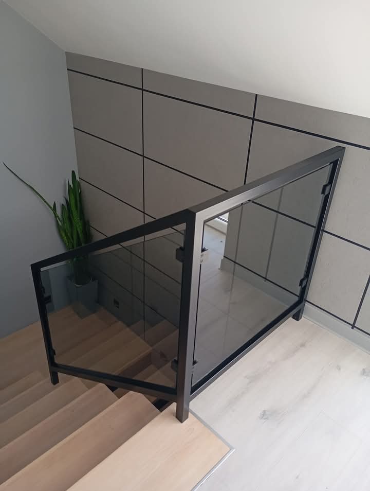 Szklana balustrada schodowa z czarną ramą, widok z góry na drewniane schody i minimalistyczne wnętrze z rośliną doniczkową w tle.