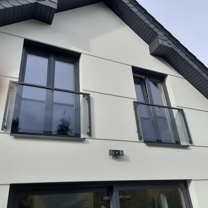 Dwa okna balkonowe z szarymi ramami i szklanymi balustradami na elewacji domu o jasnej strukturze.