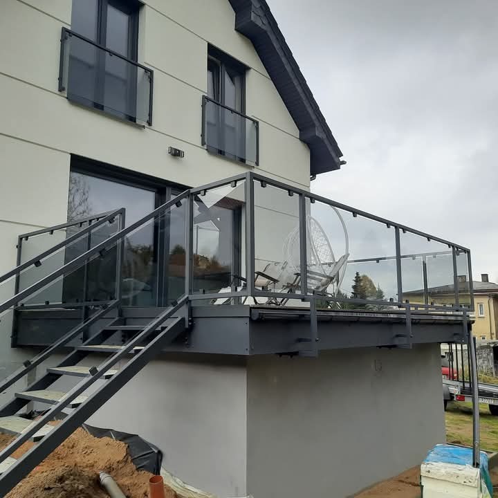 Szklana balustrada z metalowymi elementami na tarasie domu jednorodzinnego, widoczne schody wejściowe i meble ogrodowe, pochmurne niebo w tle.