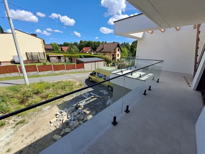 Nowoczesna balustrada nierdzewna z czarnymi słupkami i szklanymi panelami na tarasie domu jednorodzinnego z widokiem na okolicę, gdzie w tle zaparkowany jest żółty bus.