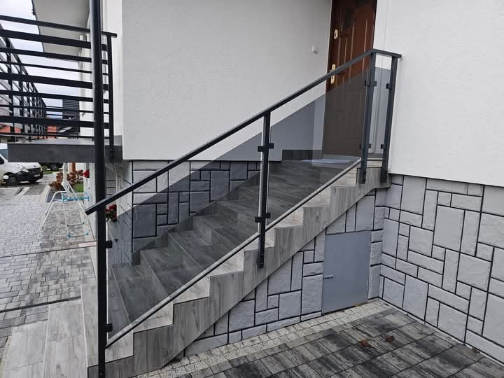 Zewnętrzne schody z szarymi płytkami, zabezpieczone czarną balustradą z elementami szklanymi, prowadzące do drzwi wejściowych.