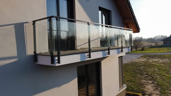 Szklana balustrada balkonowa z czarnymi elementami mocującymi, widok z dołu na tle krajobrazu wiejskiego.