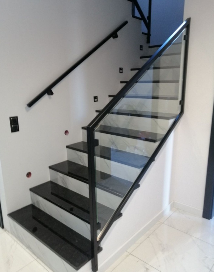 Schody z czarnego granitu z białymi wstawkami, zabezpieczone szklaną balustradą z czarnymi, metalowymi ramami i poręczą, widok z dołu.