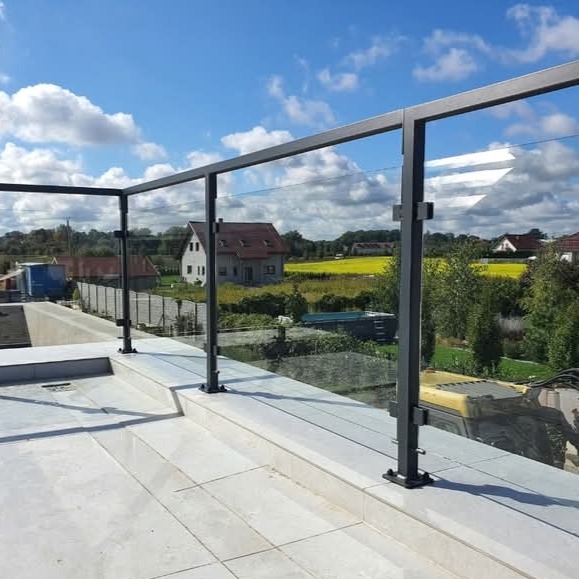 Szklana balustrada z ciemnoszarą ramą na tarasie, widok na pola i domy w tle, błękitne niebo z obłokami.
