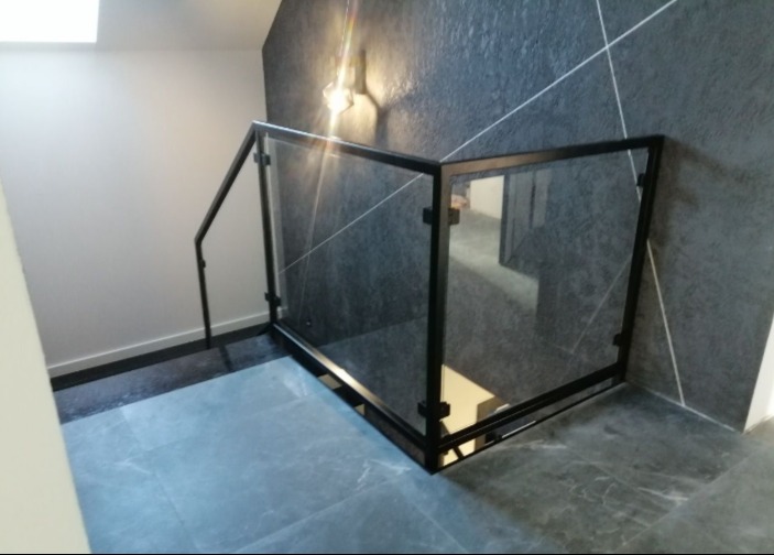 Szklana balustrada z czarną ramą na schodach z szarymi płytkami, oświetlona lampą ścienną na ciemnej ścianie z geometrycznym wzorem.