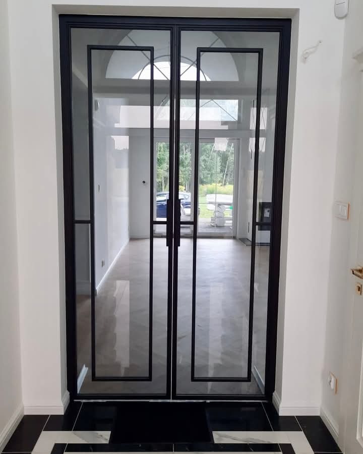 Dwuskrzydłowe szklane drzwi z czarną ramą i geometrycznym wzorem, widoczne z perspektywy wnętrza na tle jasnego pomieszczenia z dużym oknem i marmurową podłogą.