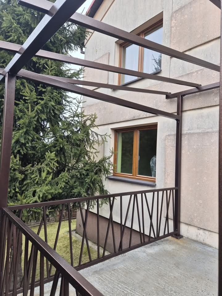 Zadaszony balkon z metalową balustradą o geometrycznym wzorze, brązowa konstrukcja na tle elewacji budynku i zielonego drzewa.