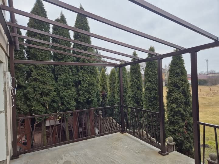 Metalowa balustrada w kolorze brązowym z prostym wzorem na tarasie, z widokiem na rząd wysokich tuj i oddalony krajobraz.