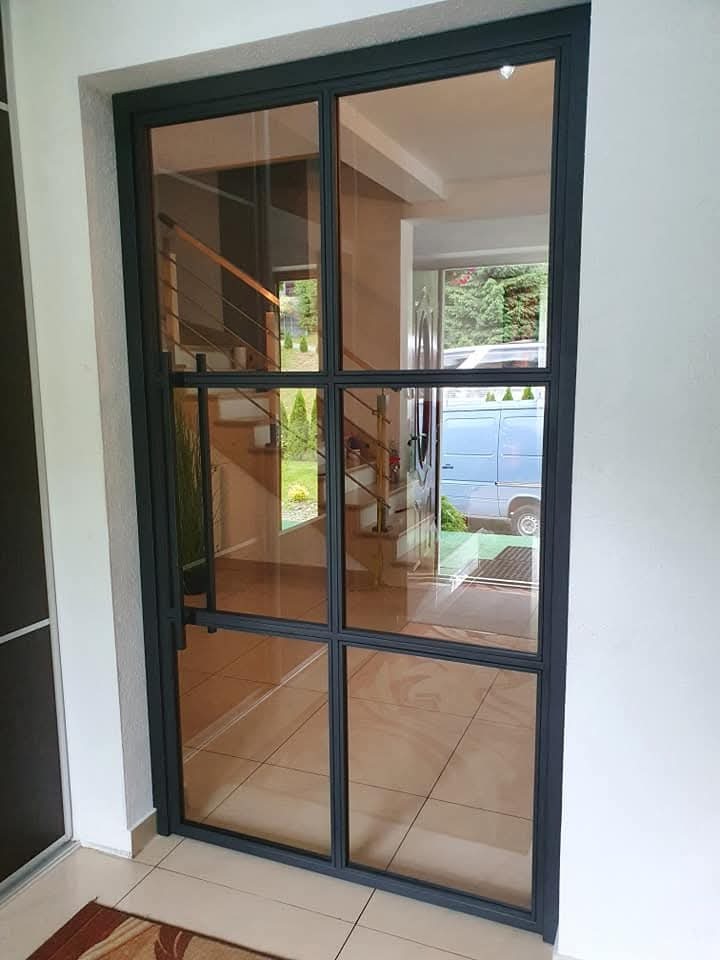 Nowoczesne, przeszklone drzwi wejściowe z czarnymi szprosami, odbijające wnętrze domu z drewnianymi schodami i zewnętrzny ogród z zaparkowanym niebieskim vanem.