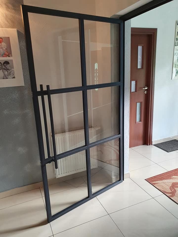 Szklane drzwi loftowe z czarną ramą, widoczne detale konstrukcyjne i uchwyt, na tle jasnej podłogi i fragmentu ściany z dekoracją.