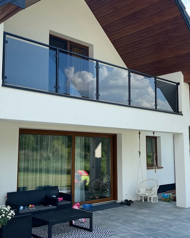 Nowoczesny balkon z czarną, szklaną barierką, odbijającą chmury, na tle białej fasady domu z drewnianym wykończeniem dachu.