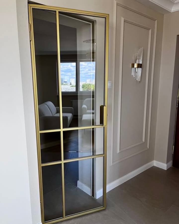 Szklane drzwi z metalową ramą w kolorze złotym, podzielone na prostokątne panele, widoczne na tle jasnego, nowocześnie urządzonego wnętrza z sofą i widokiem na miasto.