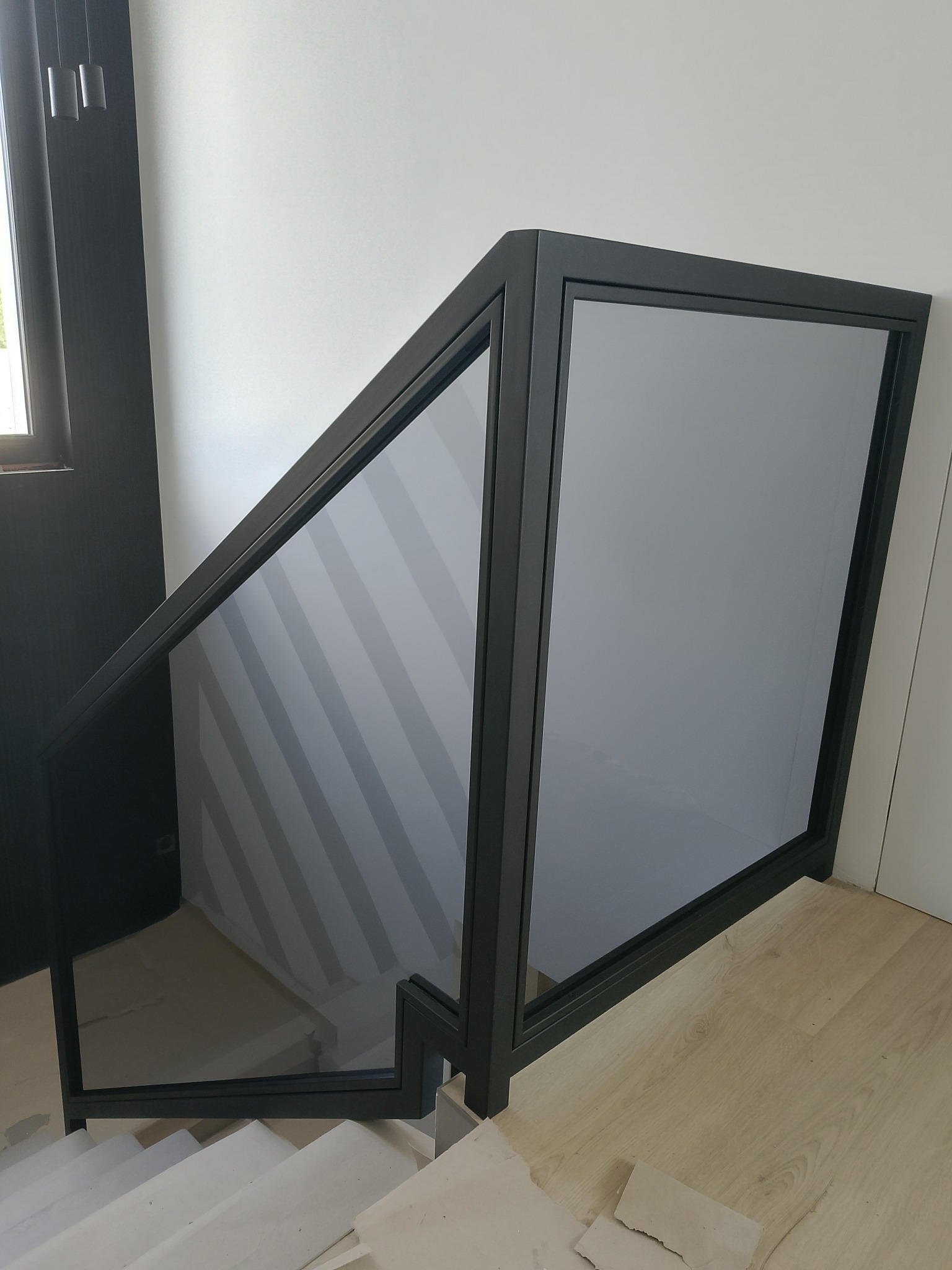 Nowoczesna balustrada szklana z czarną ramą, zamontowana na schodach wewnętrznych. Minimalistyczny design, jasne tło, widoczne schody i fragment podłogi.