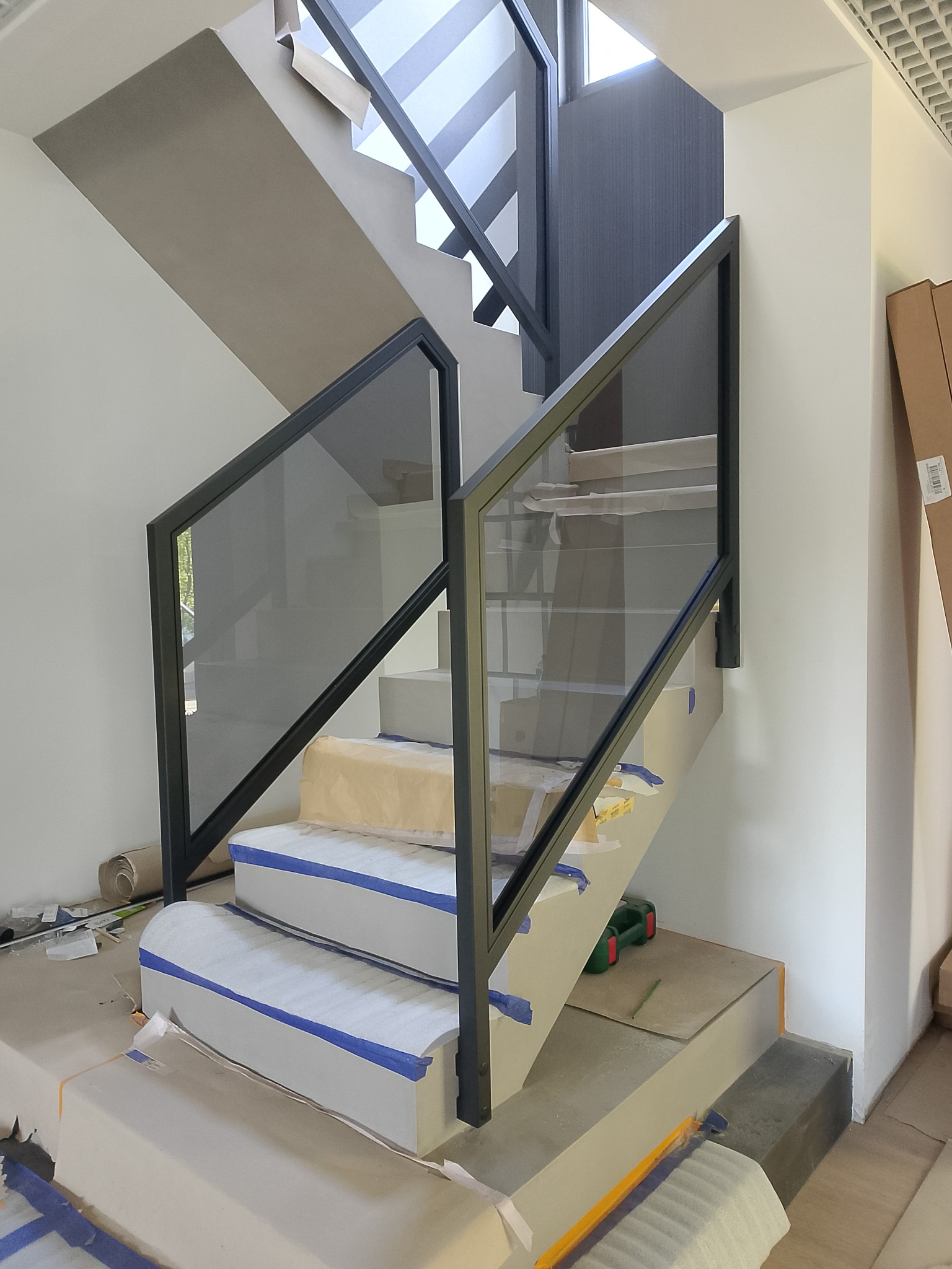 Szklane balustrady z czarnymi profilami na schodach wewnętrznych w trakcie montażu, zabezpieczone folią. Widoczne narzędzia i materiały montażowe.