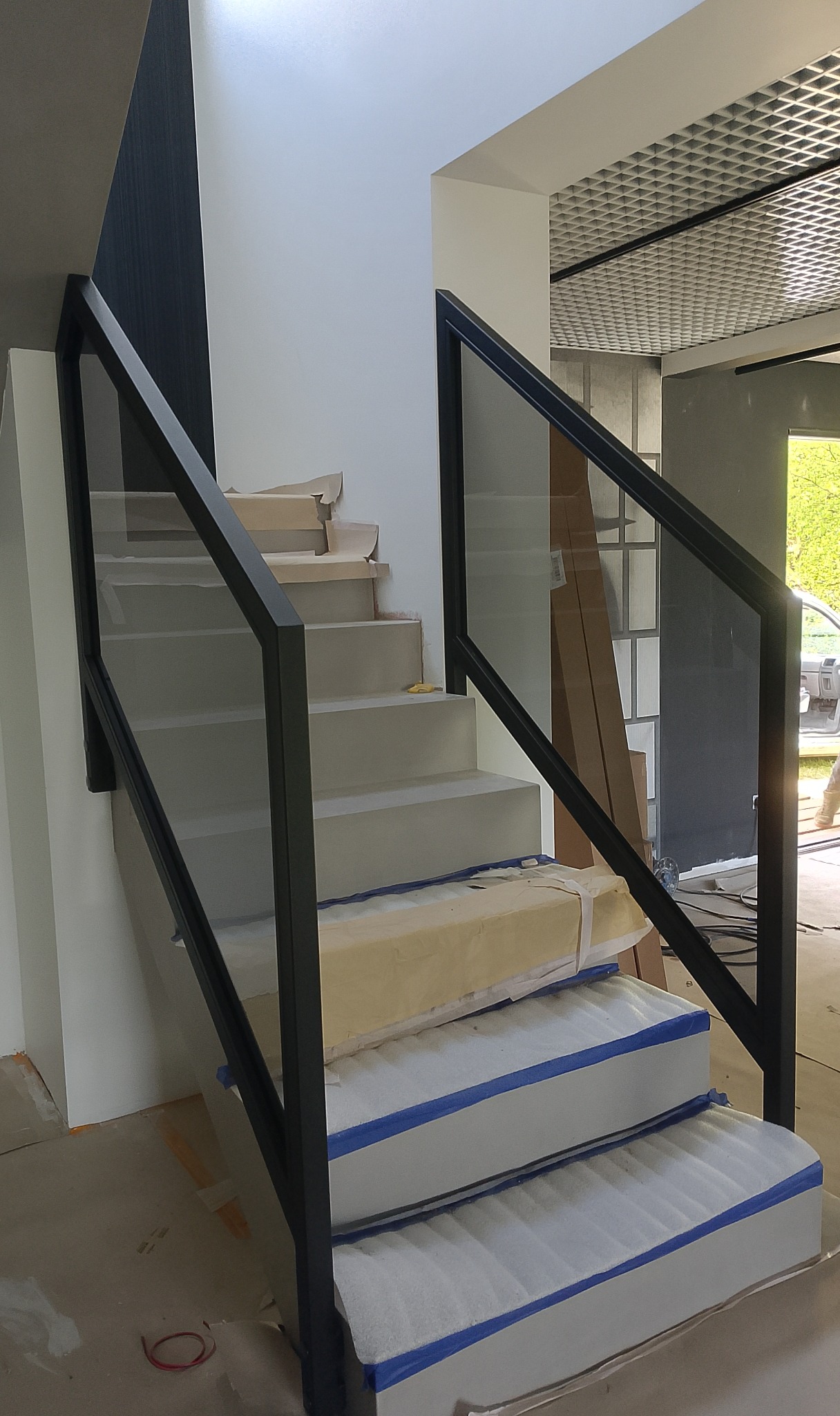 Nowoczesna balustrada szklana z czarnymi profilami na schodach w trakcie montażu. Widoczne zabezpieczenia stopni. Minimalistyczny design i jasne wnętrze.