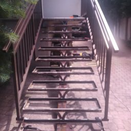 OBRA Yuriy Ruban - Metalowa konstrukcja schodów zewnętrznych w trakcie spawania, widoczne narzędzia i elementy konstrukcyjne, tło z kostki brukowej i zieleni.