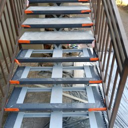 OBRA Yuriy Ruban - Stalowe schody zewnętrzne w trakcie montażu, widoczne elementy konstrukcyjne i zabezpieczenia narożników w kolorze pomarańczowym, tło z kostki brukowej i foli zabezpieczającej.