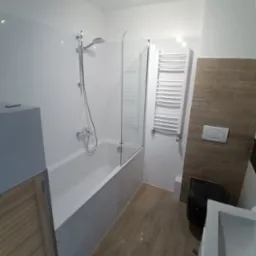 Nowoczesna łazienka po remoncie: białe płytki ścienne, wanna z szklanym parawanem, drewnopodobne płytki podłogowe, biały grzejnik drabinkowy i czarna toaleta.