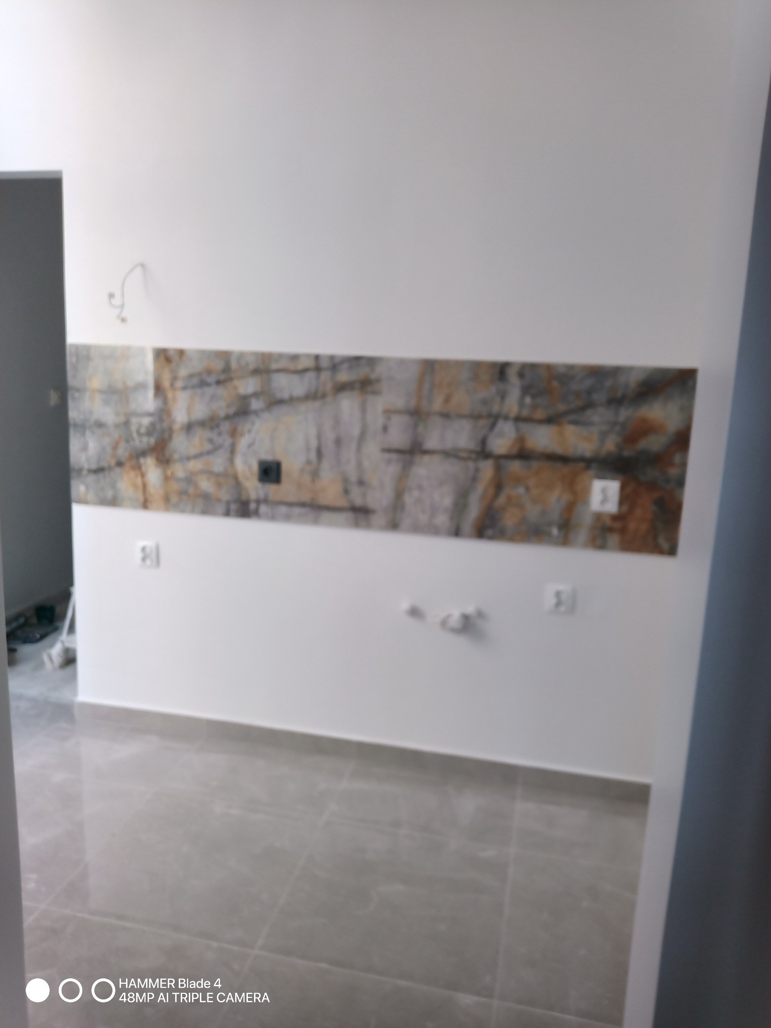 Wnętrze pokoju po remoncie z szarą płytką podłogową i dekoracyjnym panelem ściennym imitującym kamień, widoczne gniazdka elektryczne i wyprowadzone kable.