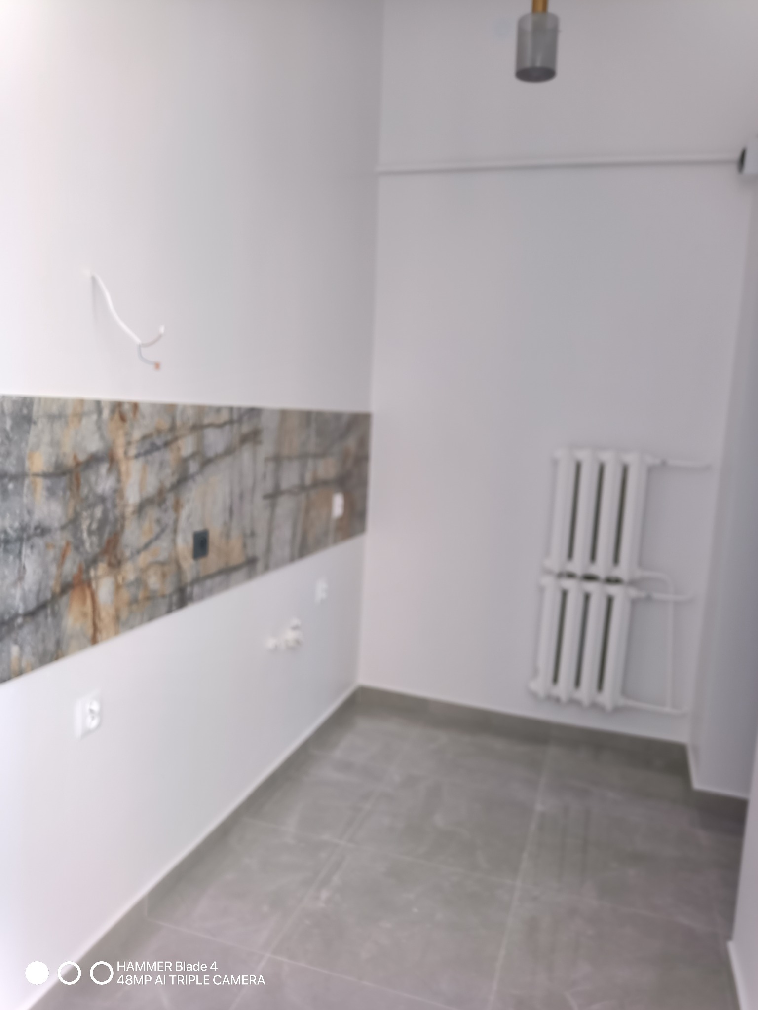 Wnętrze w trakcie wykańczania: szare płytki podłogowe, białe ściany z poziomym pasem dekoracyjnym imitującym kamień, grzejnik żeberkowy, wystające kable ze ściany.