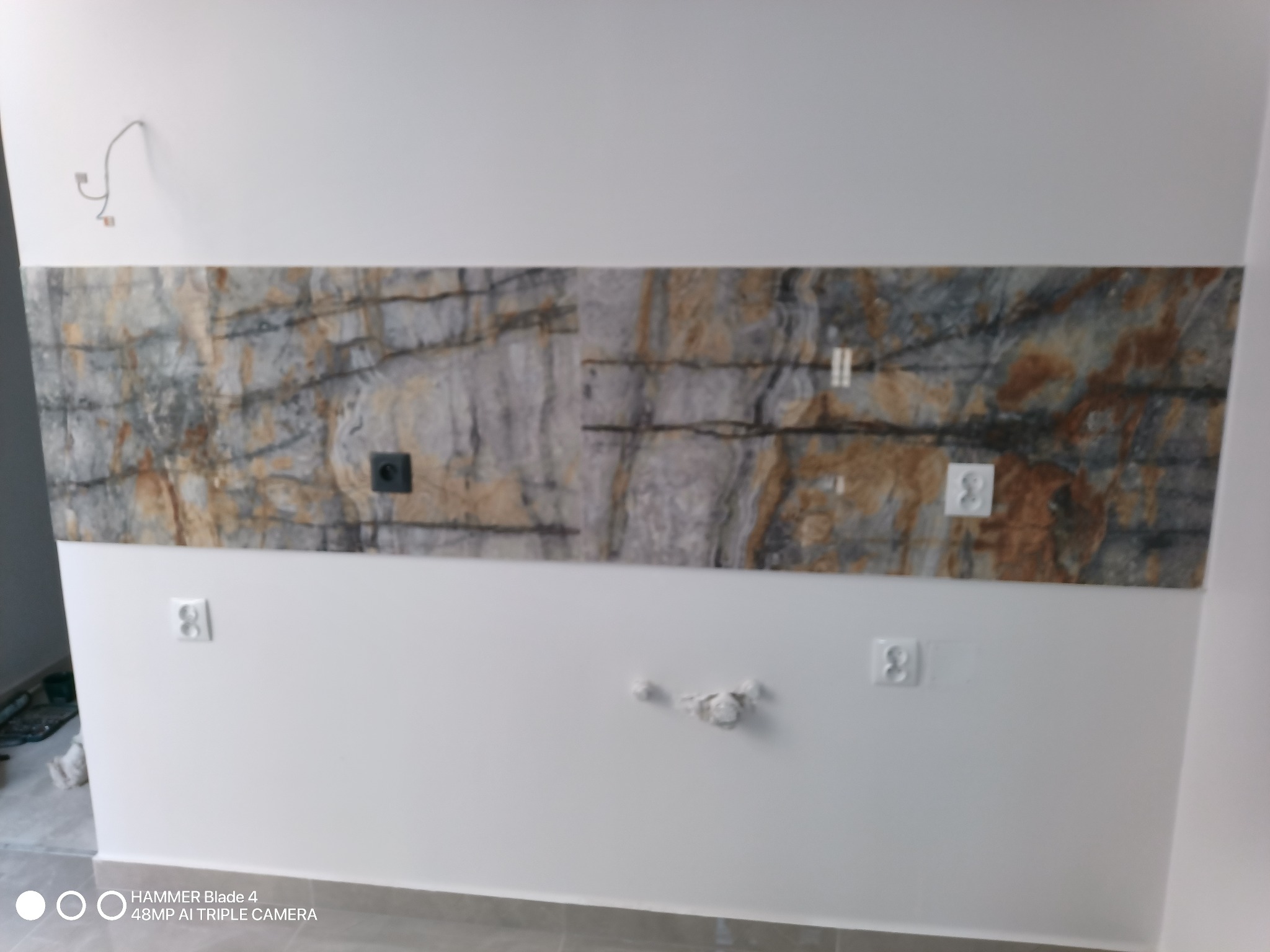 Ściana z fragmentem dekoracyjnej płyty imitującej kamień, nad gniazdkami elektrycznymi, widoczne kable wystające ze ściany, szpachlowane ślady po instalacji.