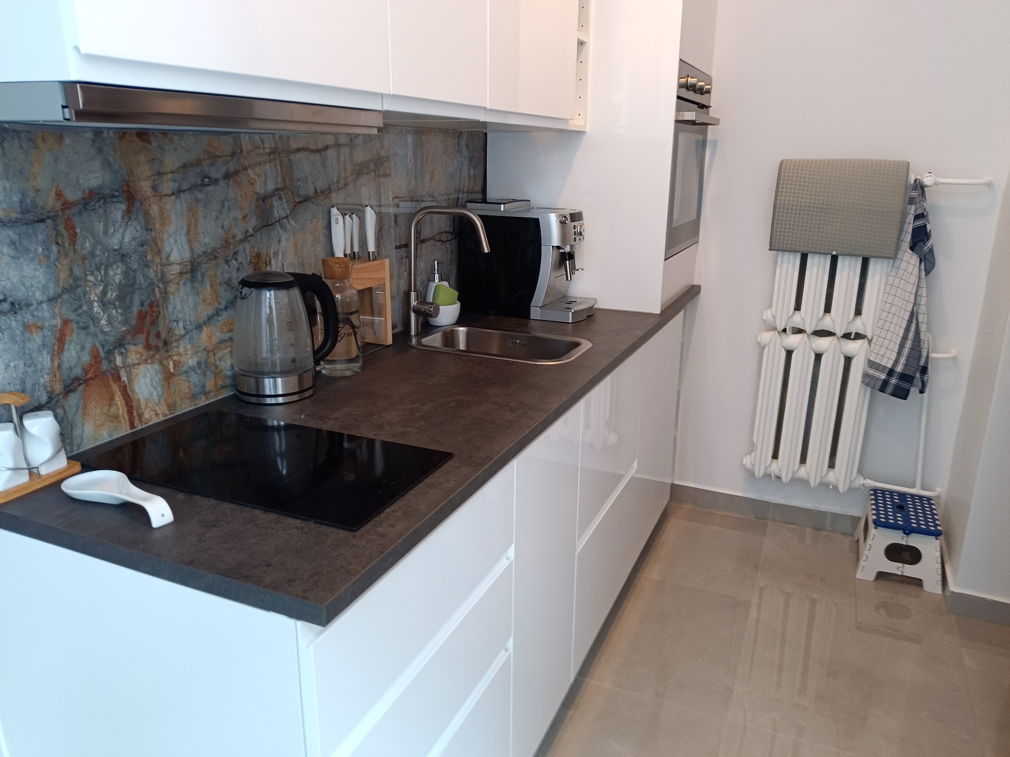 Nowoczesna kuchnia z białymi szafkami, blatem w kolorze ciemnego grafitu i marmurowym panelem ściennym, wyposażona w płytę indukcyjną, czajnik elektryczny, ekspres do kawy i biały grzejnik...