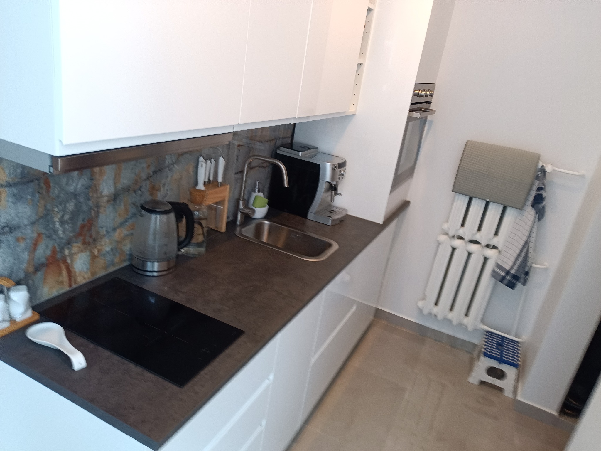 Nowoczesna, jasna kuchnia z białymi szafkami, ciemnym blatem, płytą indukcyjną, czajnikiem elektrycznym, ekspresem do kawy i grzejnikiem drabinkowym z ręcznikami.