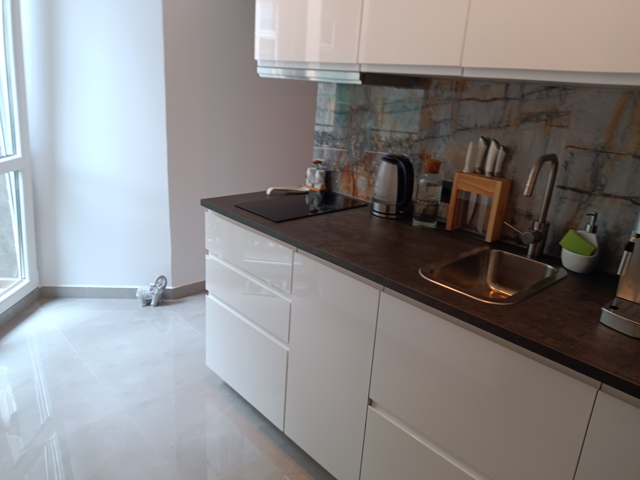 Nowoczesna kuchnia z białymi, lakierowanymi frontami szafek, blatem w kolorze ciemnego drewna i dekoracyjnym panelem ściennym imitującym kamień. Na blacie czajnik elektryczny, płyta indukcyjna...