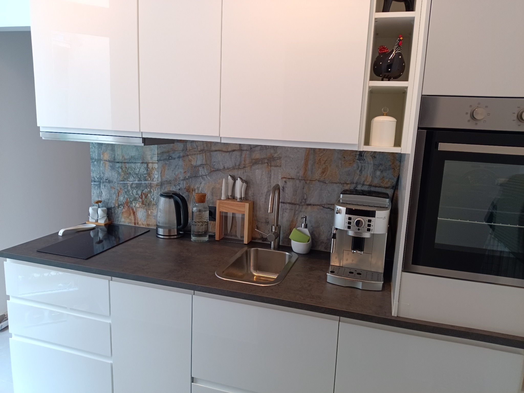 Nowoczesna kuchnia z białymi, lakierowanymi szafkami, ciemnym blatem i kamiennym panelem nad zlewem, wyposażona w płytę indukcyjną, czajnik elektryczny i ekspres do kawy.