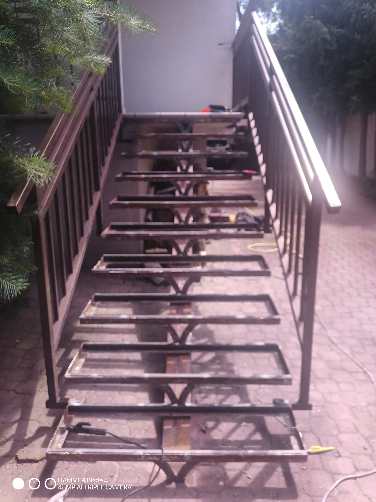 Metalowa konstrukcja schodów zewnętrznych w trakcie spawania, widoczne narzędzia i elementy konstrukcyjne, tło z kostki brukowej i zieleni.