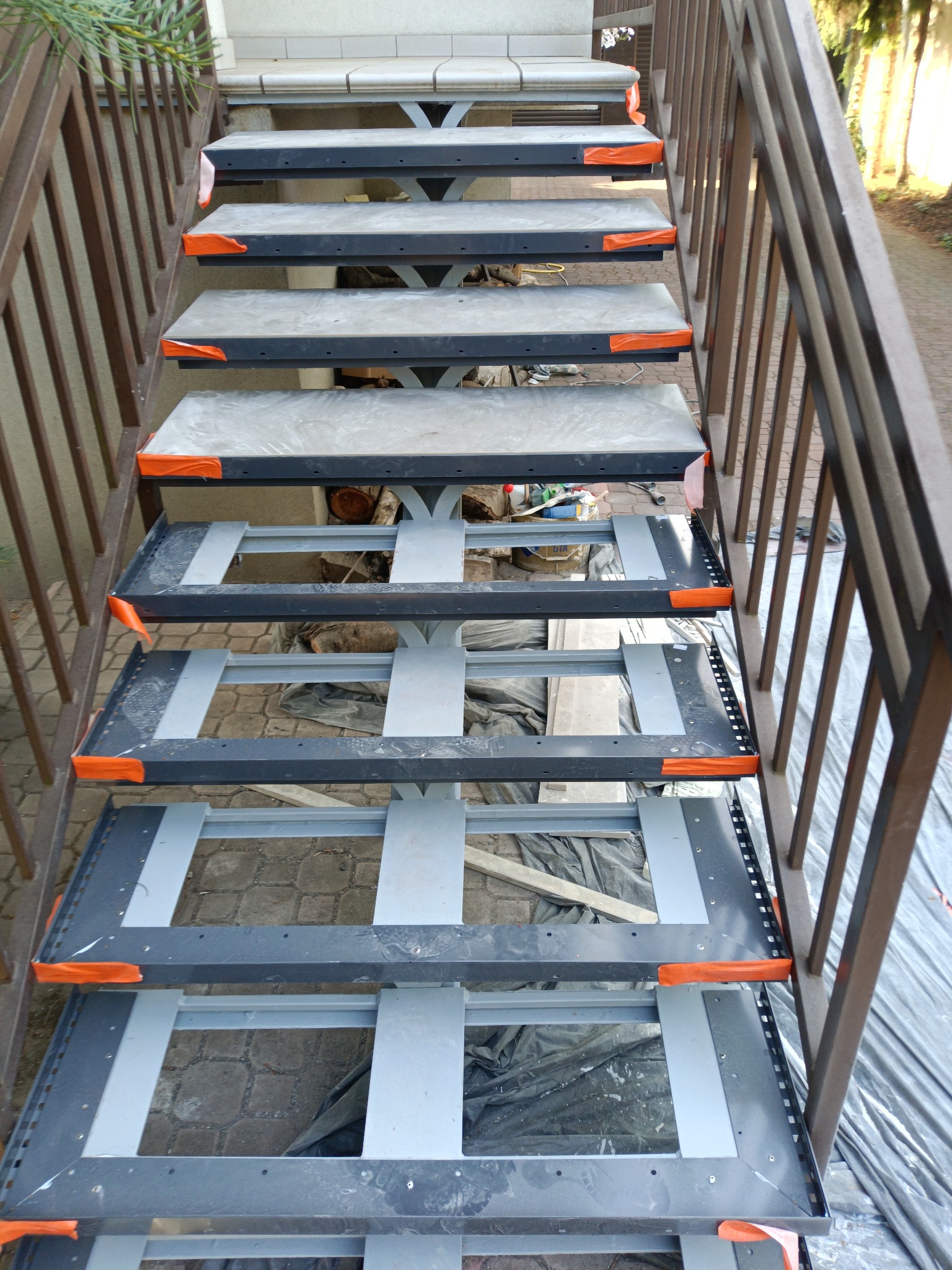 Stalowe schody zewnętrzne w trakcie montażu, widoczne elementy konstrukcyjne i zabezpieczenia narożników w kolorze pomarańczowym, tło z kostki brukowej i foli zabezpieczającej.