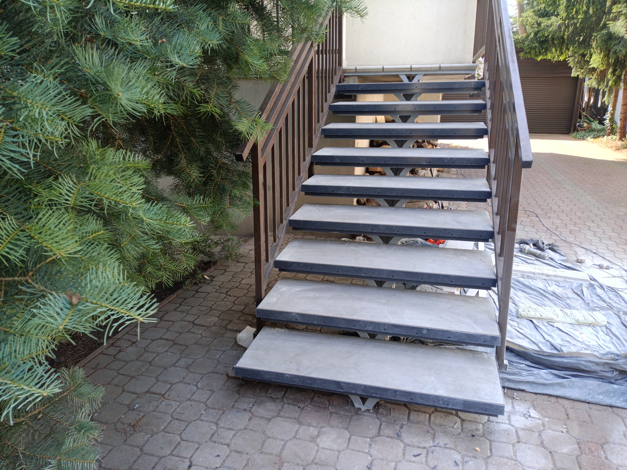 Montaż betonowych schodów zewnętrznych z brązową metalową barierką, widoczna folia ochronna i narzędzia na brukowanej powierzchni.