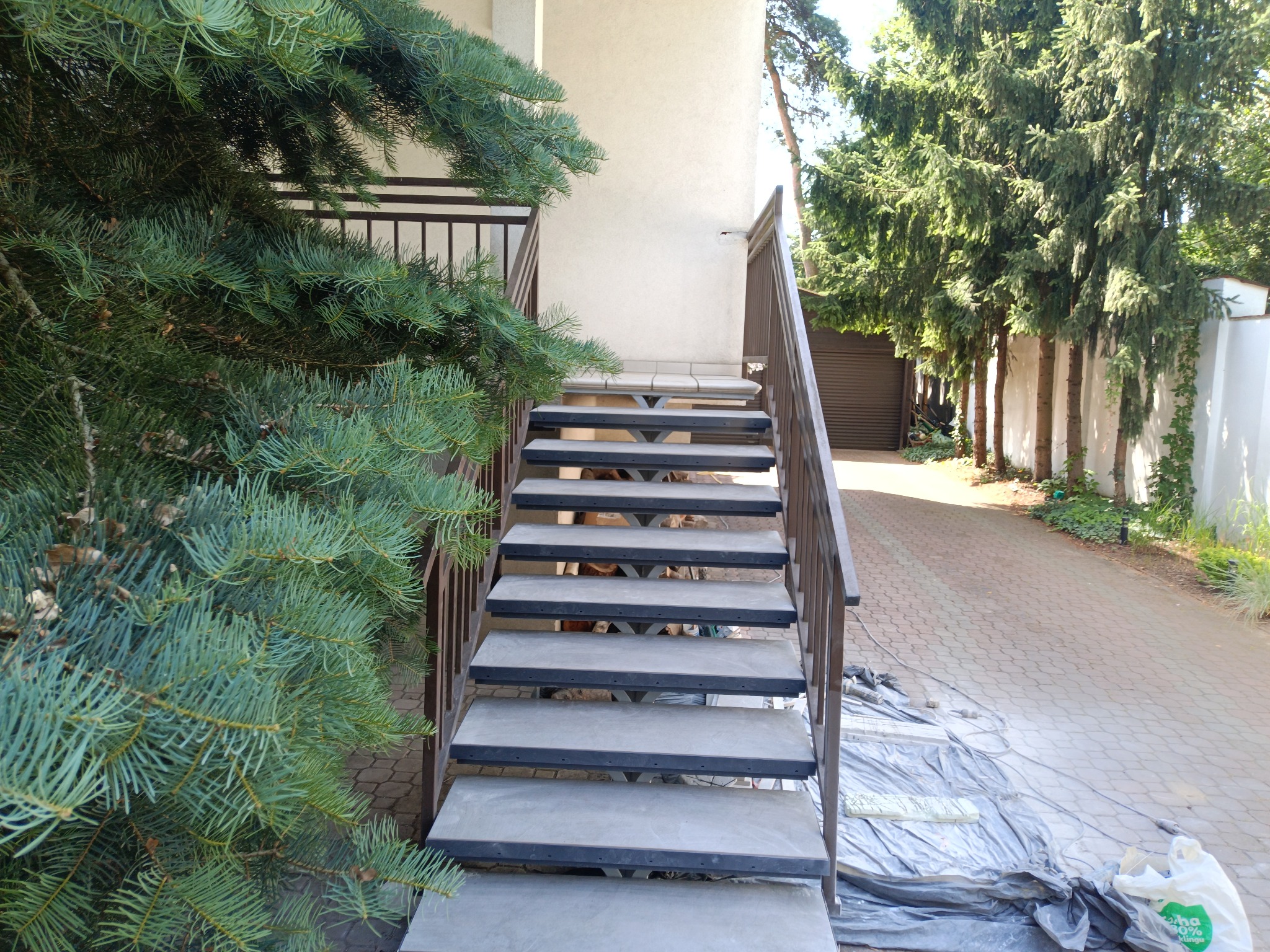 Zewnętrzne schody betonowe z metalową balustradą prowadzące do budynku, widoczne ślady prac na brukowanej powierzchni obok, iglaste drzewo po lewej stronie.