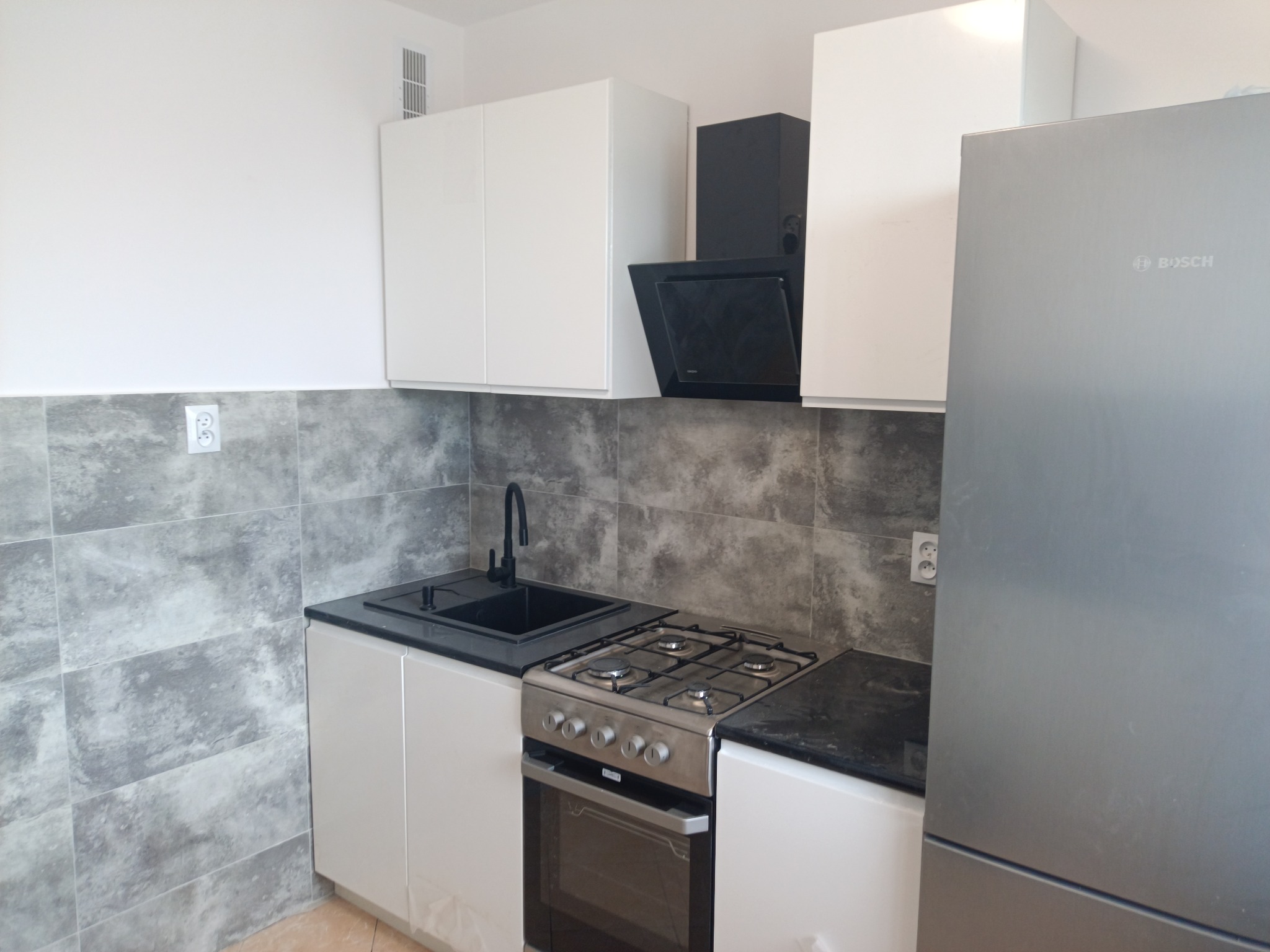 Nowoczesna kuchnia z białymi szafkami, czarnym okapem i zlewozmywakiem oraz srebrną lodówką Bosch, wykończona szarymi płytkami imitującymi kamień.