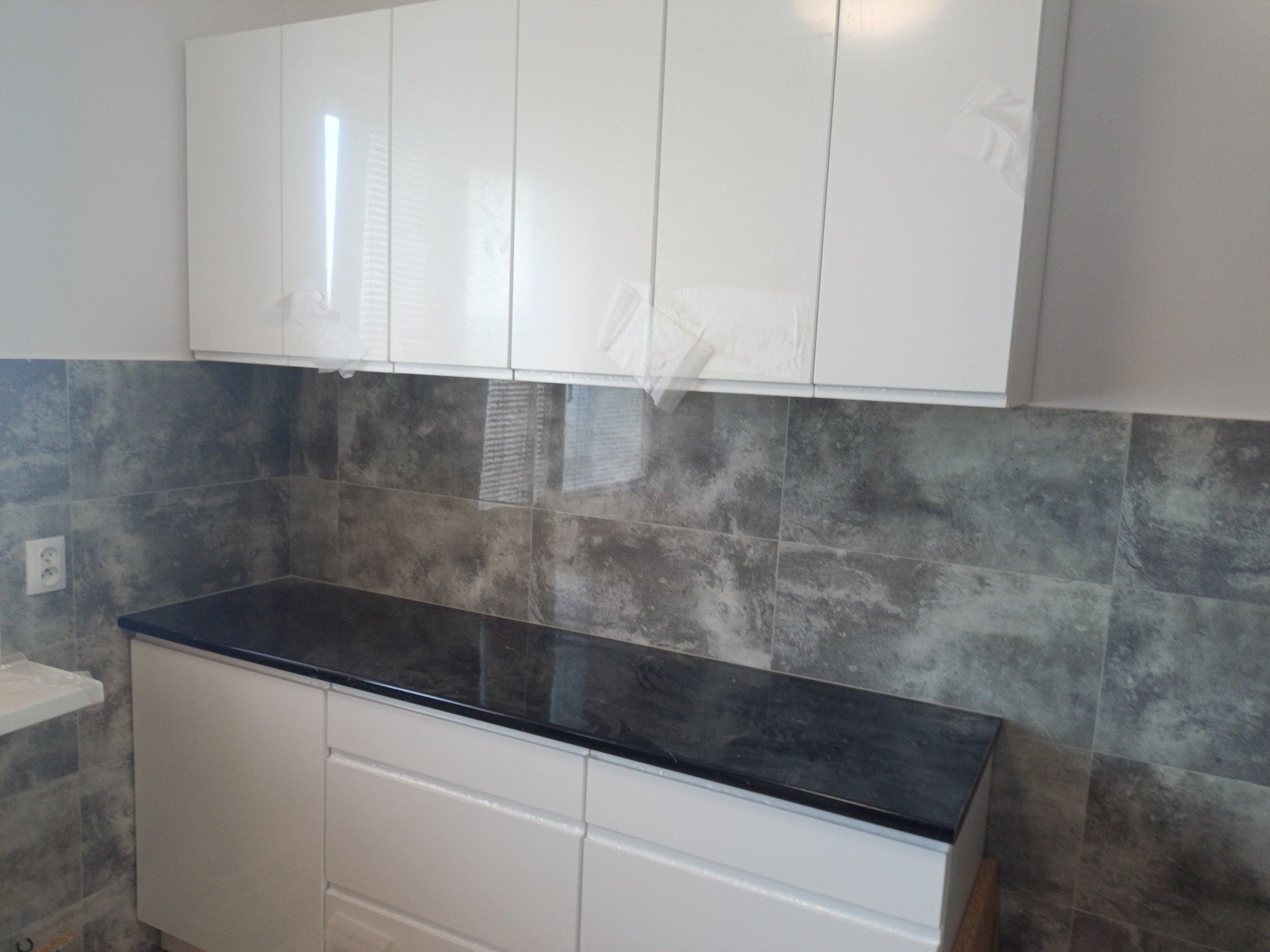 Nowoczesna kuchnia po remoncie: białe, błyszczące szafki wiszące z folią ochronną częściowo zdjętą, szare płytki na ścianie i czarny blat.