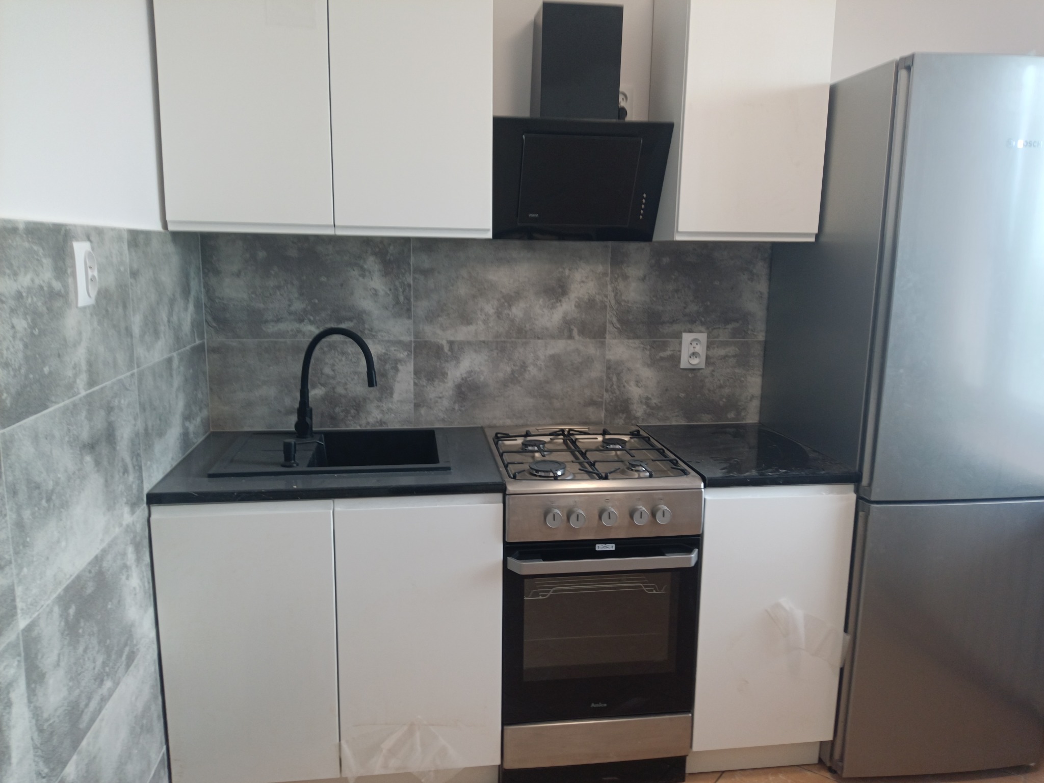 Nowoczesna kuchnia z białymi szafkami, czarnym blatem i szarą glazurą imitującą kamień, z widocznym zlewem, kuchenką gazową i lodówką.