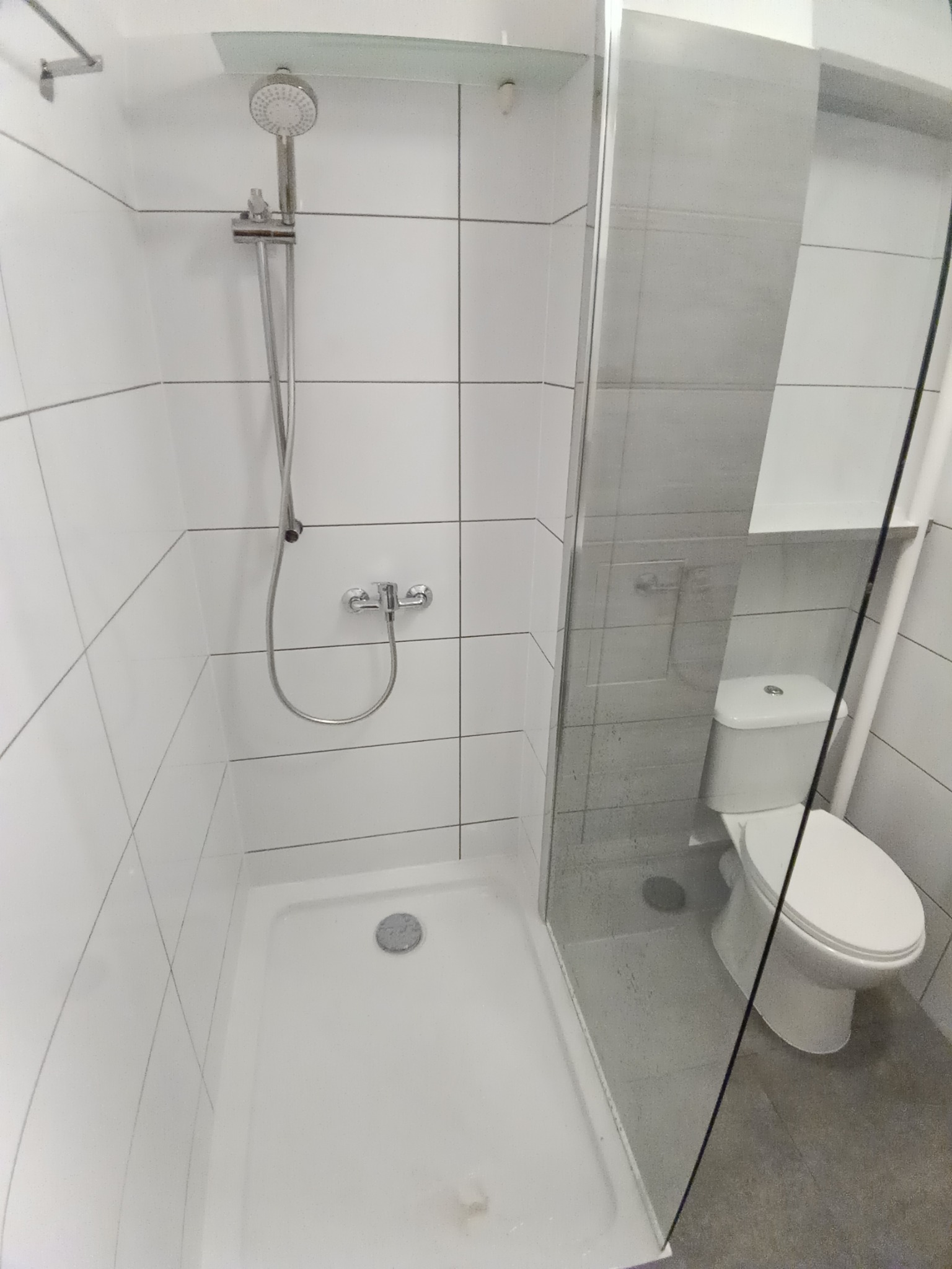 Nowoczesna kabina prysznicowa z białymi płytkami i szklanymi drzwiami, widoczny brodzik z odpływem oraz toaleta w tle, minimalistyczny design.