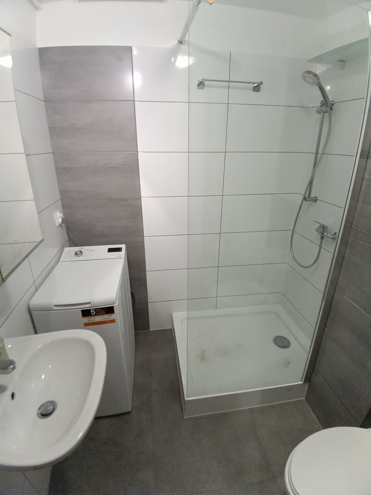 Nowoczesna łazienka z kabiną prysznicową typu walk-in, pralką obok umywalki i szarymi płytkami częściowo na ścianach.