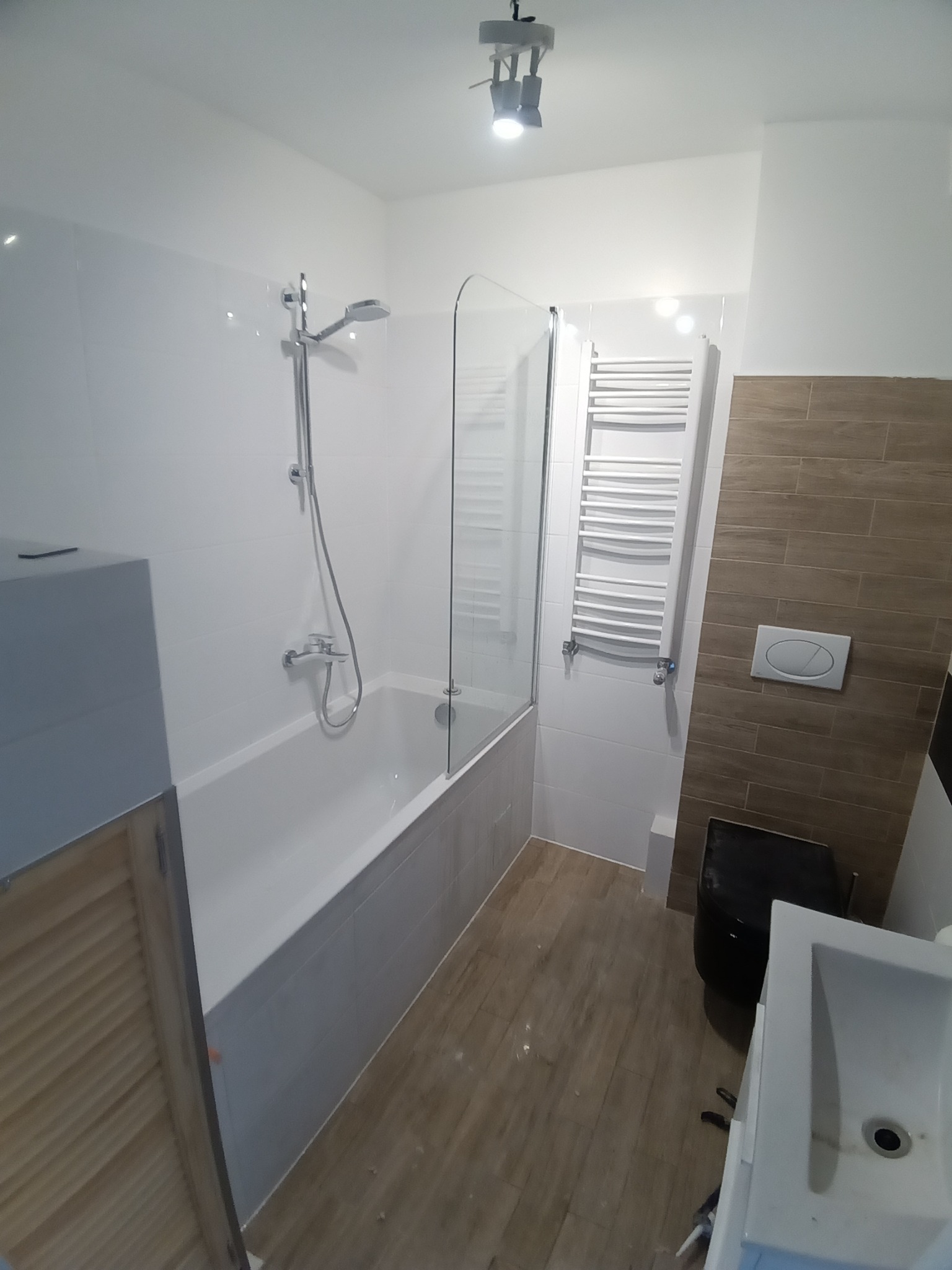 Nowoczesna łazienka po remoncie: białe płytki ścienne, wanna z szklanym parawanem, drewnopodobne płytki podłogowe, biały grzejnik drabinkowy i czarna toaleta.