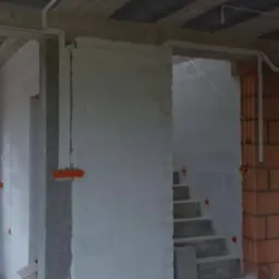 Wnętrze w stanie surowym: widoczne cegły, betonowy strop, otynkowane ściany, instalacja elektryczna w peszlach i puszki elektryczne, schody betonowe, okno z widokiem na zieleń.