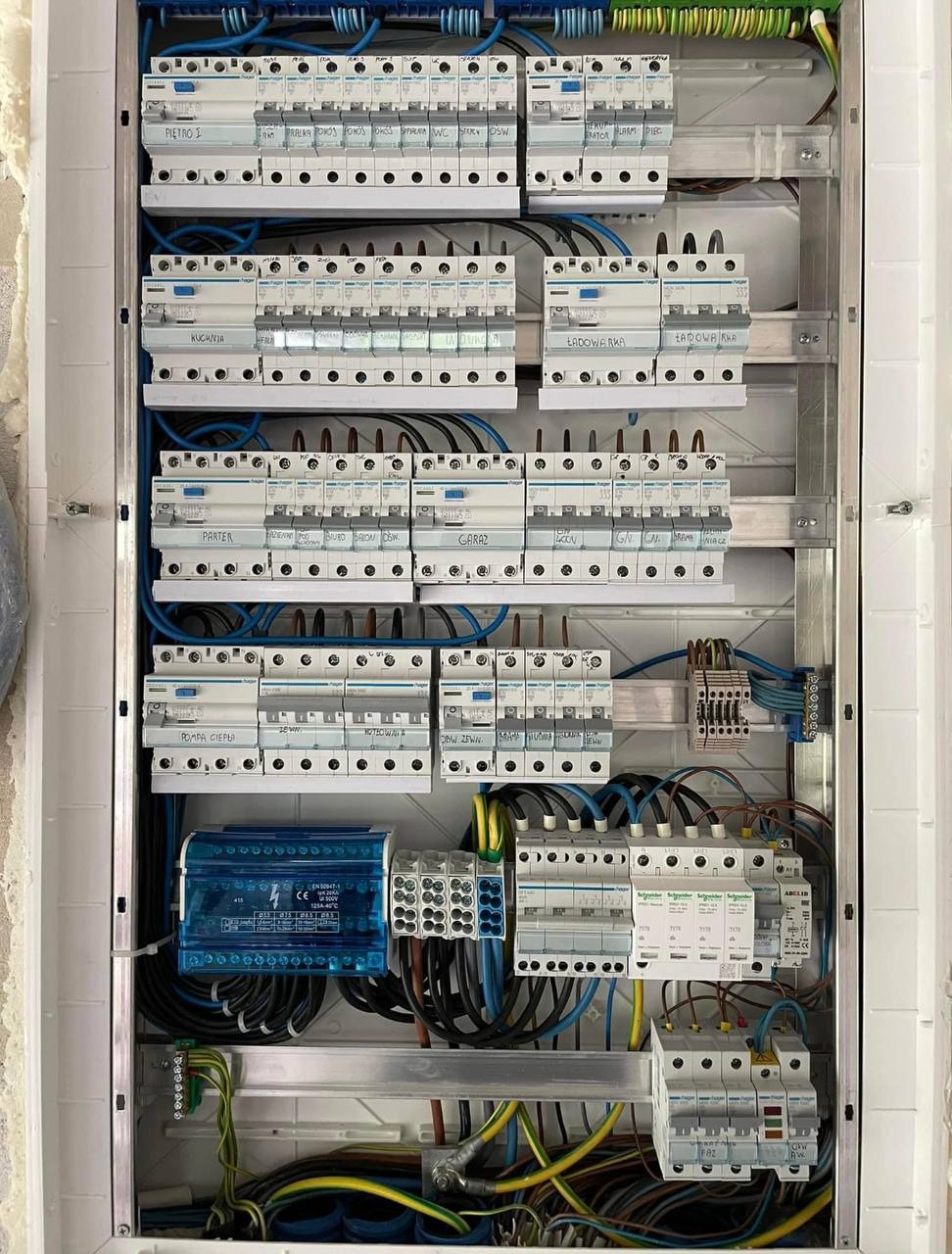 Rozdzielnica elektryczna z zainstalowanymi bezpiecznikami i okablowaniem, z opisami obwodów takimi jak 'Piętro I', 'Kuchnia', 'Parter', 'Ładowarka', widoczne przewody w różnych kolorach.