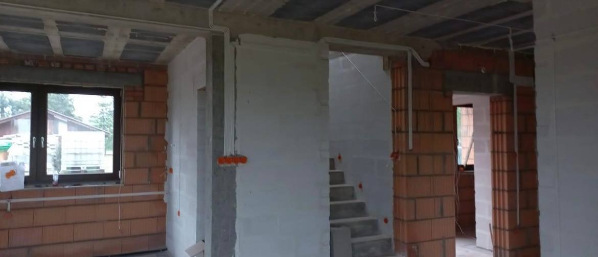 Wnętrze w stanie surowym: widoczne cegły, betonowy strop, otynkowane ściany, instalacja elektryczna w peszlach i puszki elektryczne, schody betonowe, okno z widokiem na zieleń.