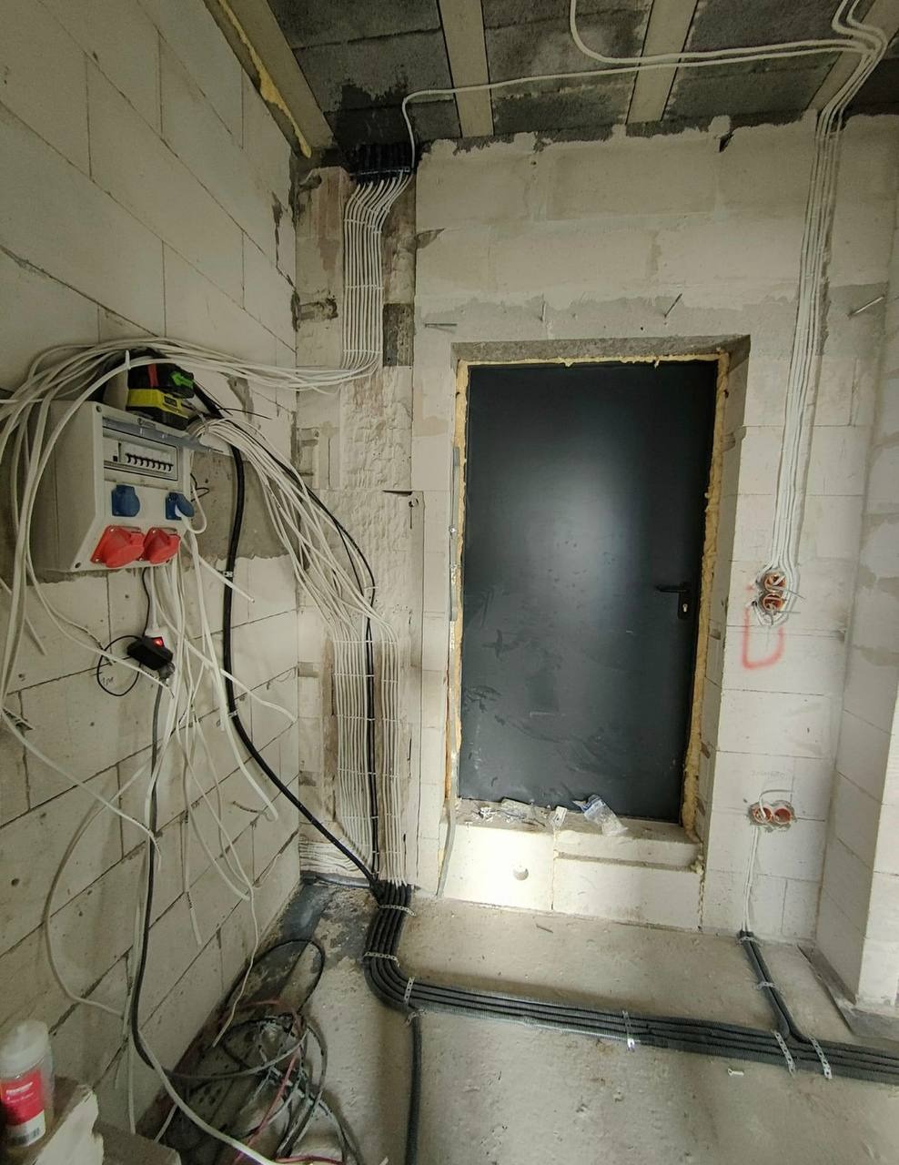 Surowy stan pomieszczenia z widoczną instalacją elektryczną: białe przewody prowadzone po ścianach z betonu komórkowego, szara skrzynka rozdzielcza z bezpiecznikami, peszle w podłodze, czarne drzwi.