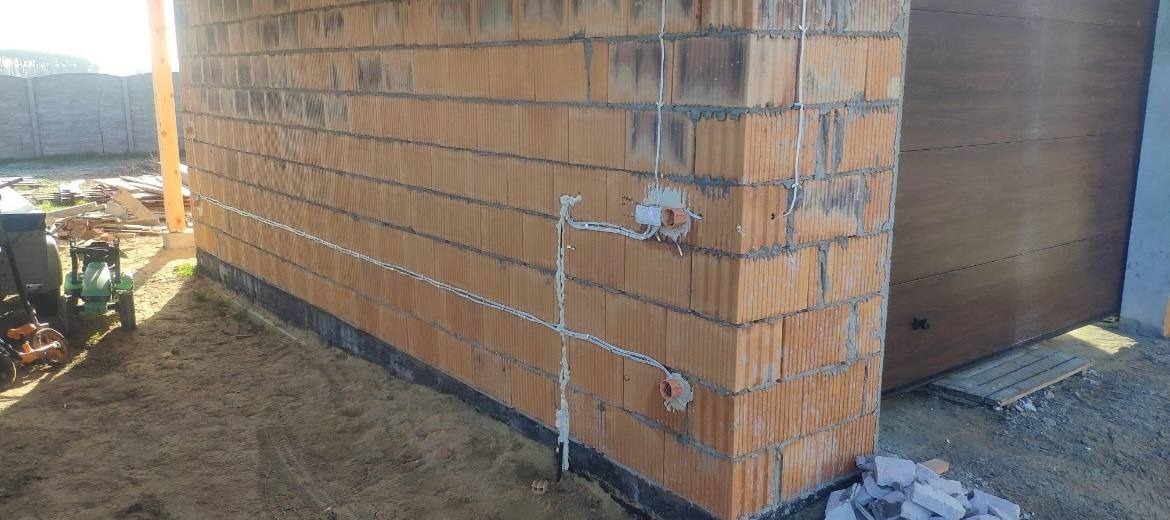 Murowana ściana budynku w trakcie budowy z widocznymi puszkami i przewodami elektrycznymi, położonymi na zewnątrz ściany.