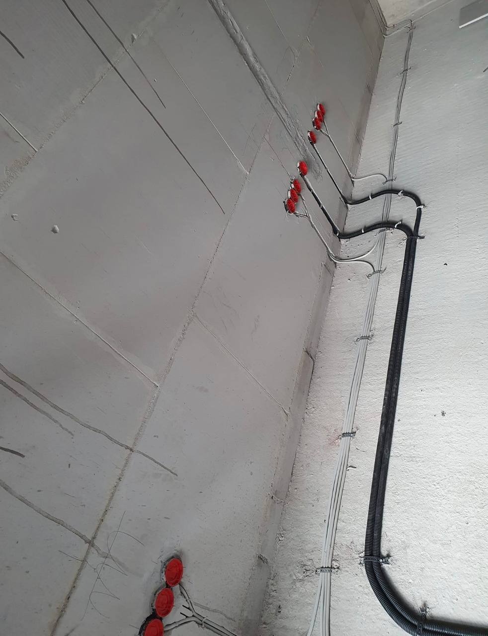 Surowa ściana z widoczną instalacją elektryczną: rurki karbowane i przewody zabezpieczone czerwonymi puszkami, w trakcie montażu.